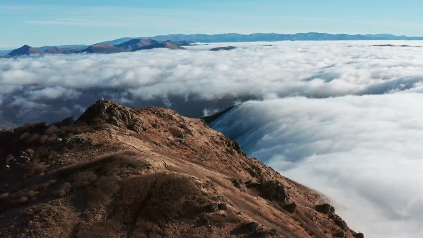 Mountain peak towering above sea of clouds under clear blue sky, solitary figure in distance Royalty-Free Stock Footage #3447672299