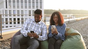 Two African American businesspeople, man and woman sitting on lunch break on beanbags at rooftop of office, looking at mobile phone sharing fun information. - Powered by Shutterstock - Get 15% off with code: PIKWIZARD15
