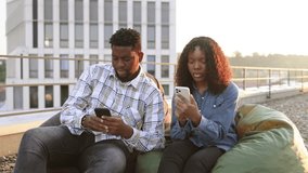 Two African American businesspeople, man and woman sitting on lunch break on beanbags at rooftop of office, looking at mobile phone sharing fun information. - Powered by Shutterstock - Get 15% off with code: PIKWIZARD15
