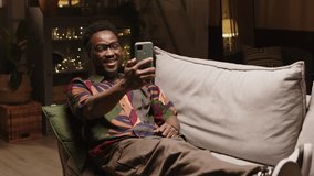 Young hipster Black man sitting in relaxed position on couch in modern living room with cozy yellow dimmed light and video calling on smartphone with mate or girlfriend, spending evening alone at home