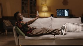 Side view of relaxed Black man in hipster clothes sitting with his feet on sofa in modern living room with yellow dimmed light and video chatting on smartphone with friend