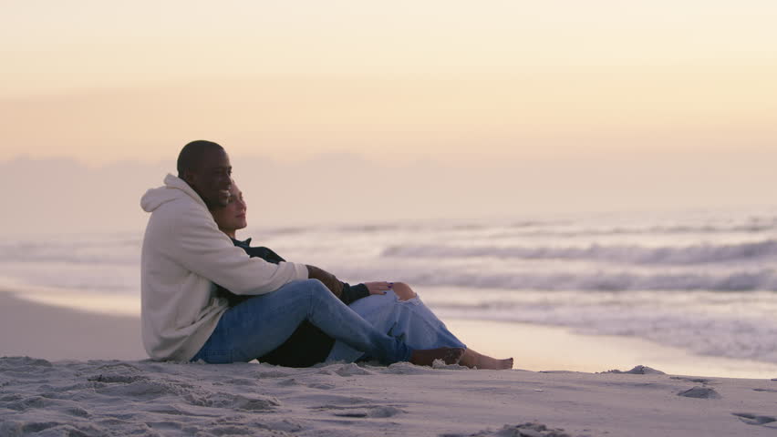 Shot of casually dressed loving young couple sitting on sand watching beautiful sunrise morning over beach and sea in South Africa - shot in slow motion