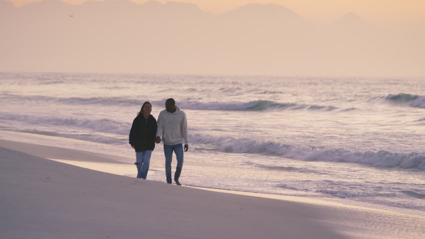 Full length shot of casually dressed loving young couple walking hand in hand along sandy shoreline watching beautiful sunrise morning over beach and sea in South Africa - shot in slow motion