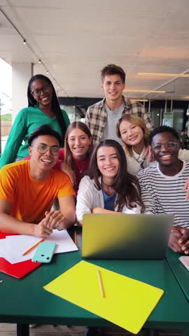 Vertical portrait. Group of young teenage students waving hands at library, using a laptop, looking at camera. Happy classmates studying and doing homeworks at highschool. Multiracial academy people