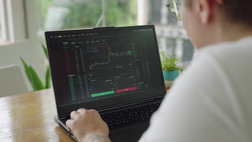 Focused individual monitors market charts on laptop in sunlit office. Analyzing real-time trading data, considering investment strategies. Professional trader engaged in day trading, clicking mouse.