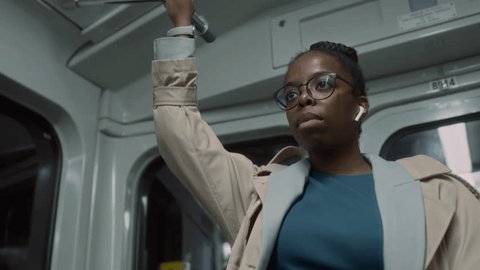 Chest up handheld footage of Black businesswoman in earbuds riding public metro train standing and holding onto handgrip while being late to meeting and checking time on watch Stock Video
