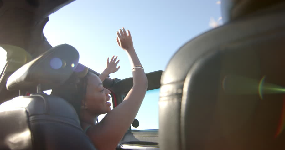 Young African American woman enjoys a sunny day in a convertible car on a road trip, with copy space. Her hands are raised in joy, capturing the essence of freedom on the road, slow motion. Royalty-Free Stock Footage #3447914859