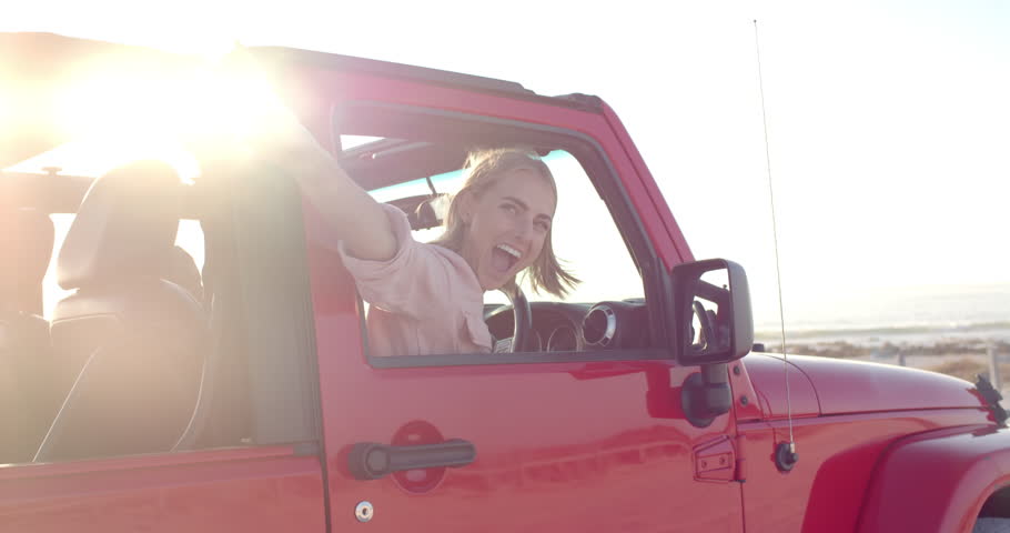 Young Caucasian woman enjoys a sunny beach drive in a red jeep on a road trip. Her cheerful expression and peace sign capture the essence of a carefree road trip, slow motion. Royalty-Free Stock Footage #3447962719
