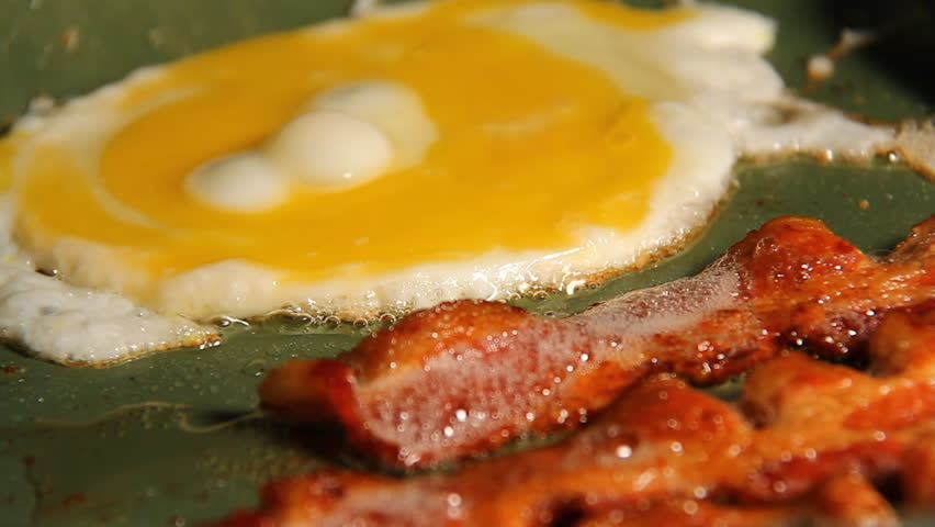 Bacon and Eggs. Three strips of bacon frying in a pan with an egg getting