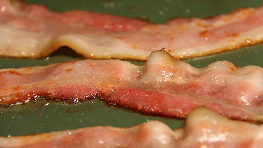 Bacon 1. Three strips of bacon frying in a pan turned over with tongs. Early in