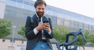 Portrait of smiling man which sitting on his bike and using his smartphone near beautiful modern city building