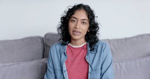 Young Asian woman sits comfortably at home on a couch on a video call. Her relaxed posture suggests a casual and cozy home environment, slow motion.