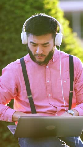 Asian man working with a laptop while using headphones in a park
