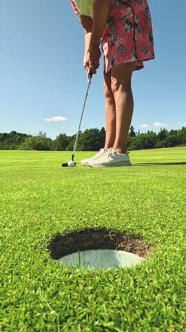 Mature woman playing golf. Golfer hitting golf shot and hammering a ball into the golf hole. Beautiful sunny landscape, with green hills and blue sky.