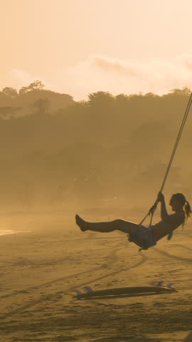 MOVING SHOT: Young couple swinging on the tropical hazy beach at sunset. Playful time on a swing on the misty beach Playa Venao. Carefree leisure activities while travelling through exotic Panama. Royalty-Free Stock Footage #3448203467
