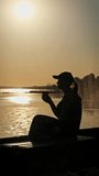 Vertical video Young woman drinks warm coffee at beautiful sunset, slow motion. Silhouette of girl in the setting sun sits on the parapet with coffee in hands and enjoys life. Cheerfulness, lifestyle