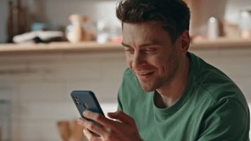 Closeup cheerful freelancer eating breakfast watching cellphone social media at home. Happy relaxed guy laughing looking funny video online at smartphone screen. Smiling man dinning with mobile phone