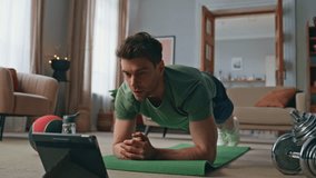 Athlete following online lesson standing plank position on yoga mat home. Sportive man doing muscle exercise looking video lesson on tablet. Active sportsman exercising with computer workout tutorial.