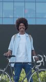 Vertical video.Smiling young african american man with afro hair in a financial district with a bicycle looking at camera 