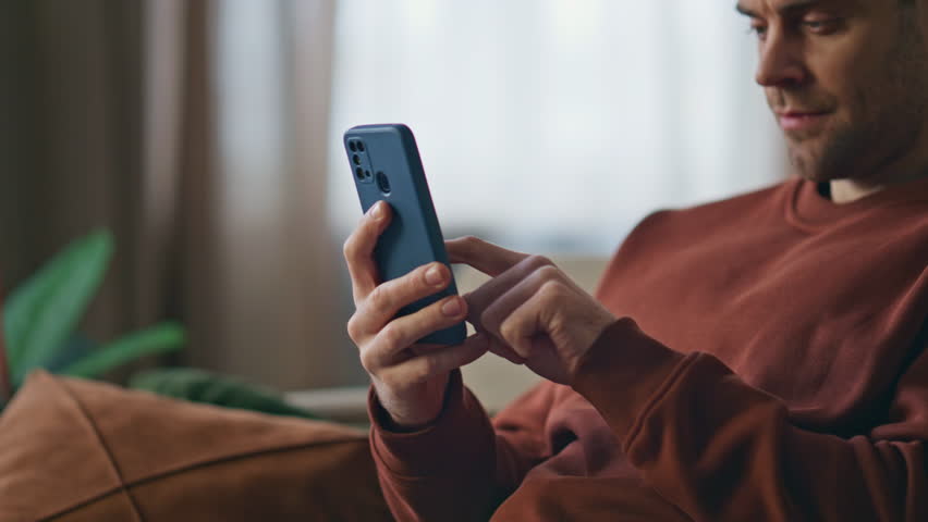 Closeup guy swiping cellphone choosing online purchase at home couch. Relaxed millennial man touching smartphone screen sitting sofa. Calm serious male scrolling news feed at mobile phone application. Royalty-Free Stock Footage #3448268345