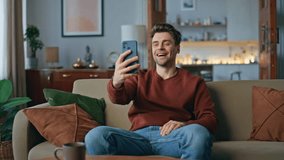 Happy blogger streaming by smartphone in modern living room. Cheerful man laughing reading live comments on cellphone screen. Positive guy influencer recording video content at mobile phone on couch.