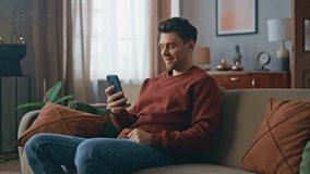 Laughing guy looking smartphone funny videos at apartment couch. Happy smiling man browsing news feed enjoying entertaining content on cellphone. Cheerful male watching social networks on mobile phone