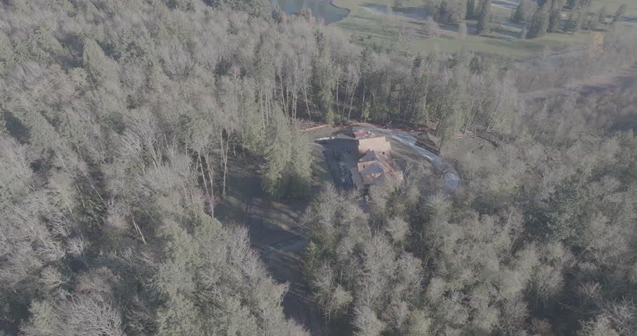 Ungraded wide-angle aerial drone footage of the wood frame of a house being built in a thick forest of evergreen and deciduous trees on a sunny winter morning in Washington State.