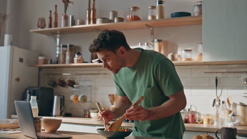 Young man watching video cooking in big kitchen closeup. Focused hipster looking laptop computer screen mixing salad in light house. Brunette guy creating vegan bowl enjoying culinary blog in flat