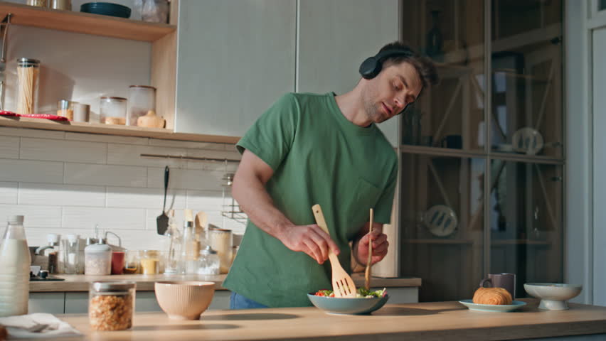 Guy in earphones preparing food dancing along favorite song at kitchen closeup. Funny joyful man wearing wireless headset listening music cooking salad with healthy vegetables. Handsome male singing