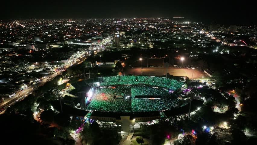 Concert of JUAN LUIS GUERRA in Olympic Stadium at night. Flashing colorful lights during open air music festival. Lighting cityscape of Santo Domingo and traffic on highway. Aerial orbit wide shot. Royalty-Free Stock Footage #3448315155