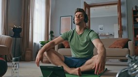 Serene athlete meditating in headphones at modern apartment. Calm fit man closing eyes sitting in lotus position at sport mat home. Relaxed young sportsman practicing zen listening tablet video lesson
