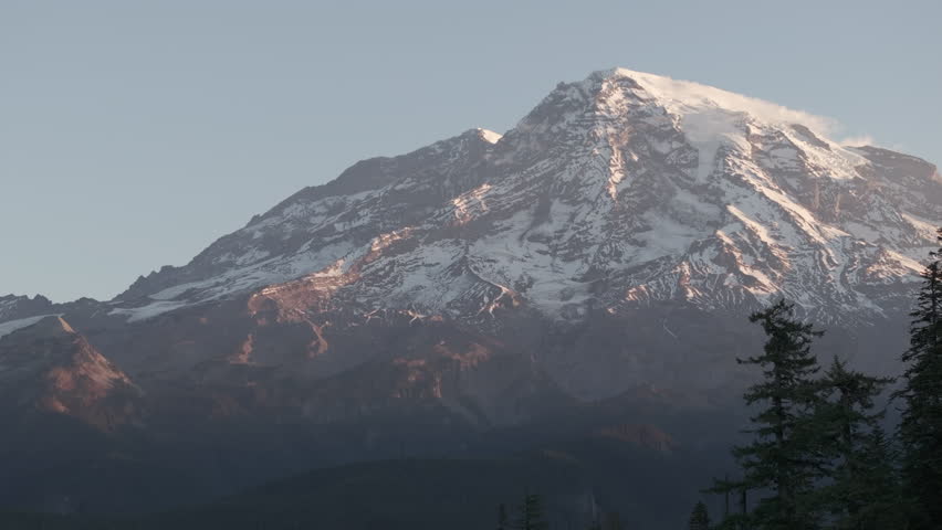 Ungraded aerial footage of Mount Rainier at dawn under light blue sky as sunrise illuminates the mountain in a soft, warm glow on a bright autumn morning in Washington State.