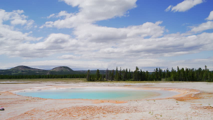 A tranquil geothermal pool mirrors the sky in Yellowstone National Park, bordered by lush forests and distant hills under a vast blue sky. Slow Motion, 4K RAW. 