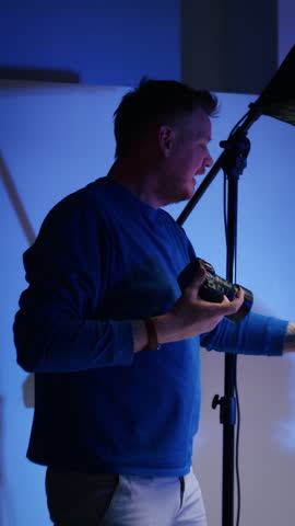 Male photographer reviewing camera in studio with blue lighting. Professional photography concept. Design for photography workshop advertising, camera equipment marketing, and editorial use. Vertical