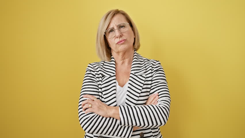 Disgruntled middle-age blonde woman in striped jacket stands crossed-armed, expressing nervous skepticism and disapproval. negatively charged personality isolated on vibrant yellow background.