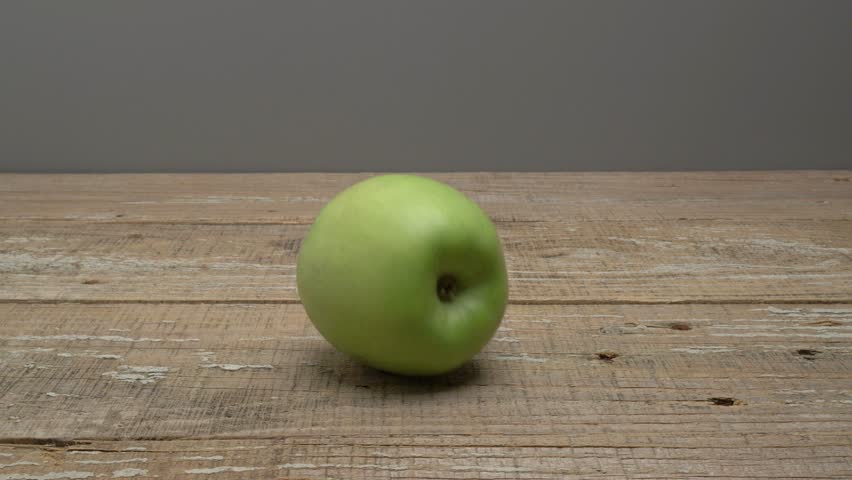 A green apple rolls on a wooden surface on a gray background.