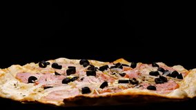 
Pizza rotates on a black background, side view, close-up. Pizza with toppings, salami, tomatoes, fresh onions, ham, olives, peppers, cheese. Fast food.