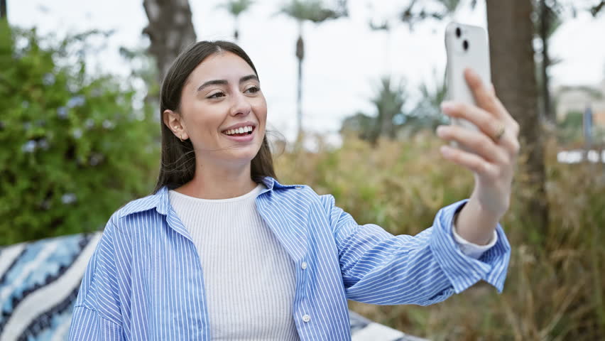Smiling young hispanic woman in a striped shirt having a video call with a smartphone in a lush outdoor park