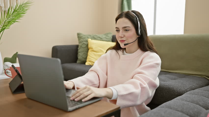 A young caucasian woman works from a cozy apartment, engaging in conversation while wearing a headset and using a laptop. Royalty-Free Stock Footage #3448768797