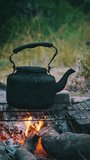 Vertical, Old kettle standing on a tourist campfire in nature. Blackened with soot smoked teapot is boiling over the self-made tourist stove made of stones in the ground. Making tea on the open fire.