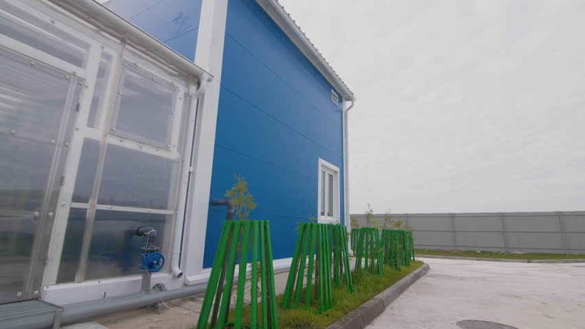 Blue industrial building with transparent wall and growing young trees in yard. Treatment plant workshop under sky with heavy clouds