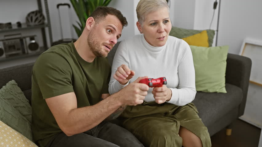 Man and woman playing with a red game controller, focused and entertained, in a cozy living room environment.