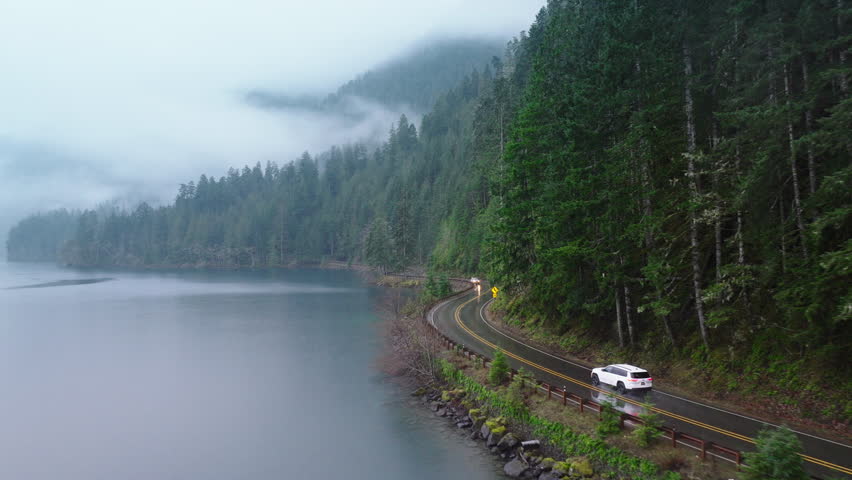 Cinematic evergreen forest Olympic National park. Travelers in rainforest Washington. Road trip concept aerial. SUV car with tourists driving by scenic route along beautiful lake Crescent on rainy day