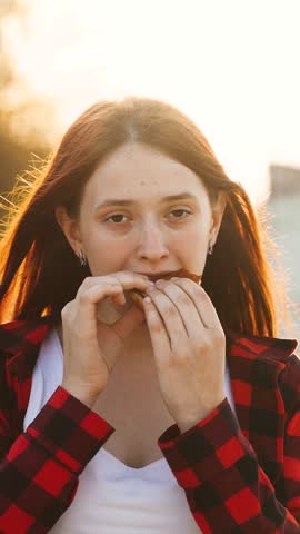 Modern young casual redhead woman eating fresh appetizing burger fast food sunny summer outdoor closeup. Confident female biting cheeseburger hamburger enjoy delicious taste at city public park