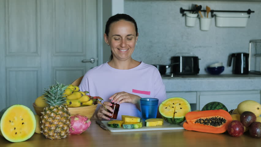 Cheerful young woman surrounded by exotic tropical organic fruits taking multivitamin pill while sitting at table, slow motion. Vitamins, nutritional supplements and healthy lifestyle concept