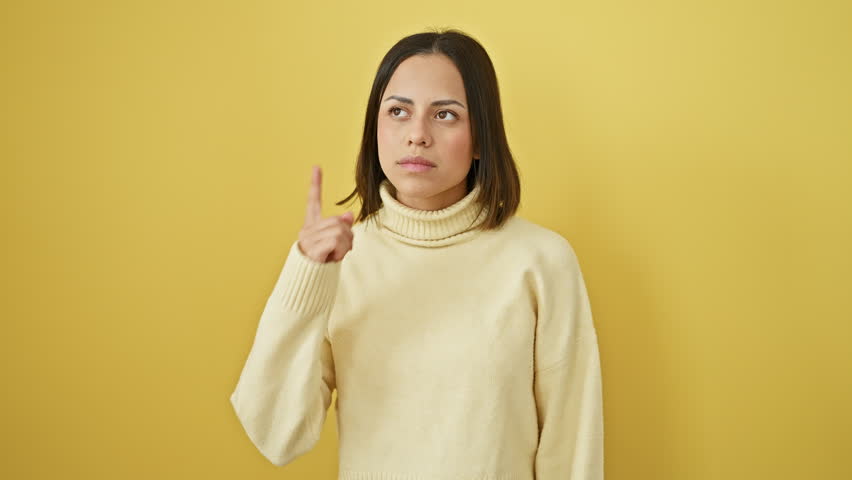 Young beautiful hispanic woman wearing sweater standing pointing finger up with successful idea. exited and happy. number one. over isolated yellow background