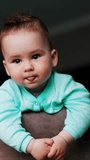 Lovely toddler looking around standing peacefully on the chair. Adorable baby showing tongue and smiling. Low angle view. Vertical video.