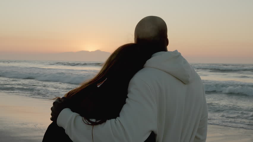 Rear view of casually dressed loving young couple watching beautiful sunrise morning over beach and sea in South Africa with lens flare - shot in slow motion