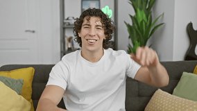 Smiling young hispanic man with curly hair wearing a white shirt, sitting on a sofa in a cozy home interior.
