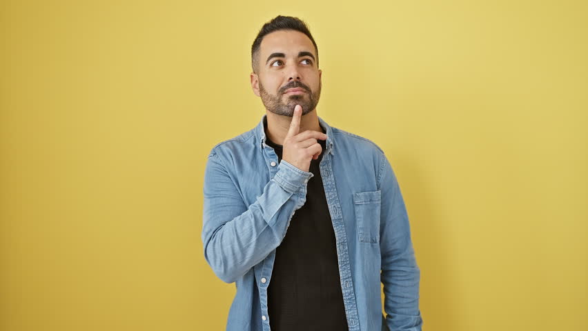 Pensive young hispanic man in denim shirt, finger on chin, lost in deep thought. looking up with a puzzled, serious expression on yellow isolated background, subtly wrestling with doubt. Royalty-Free Stock Footage #3449056513